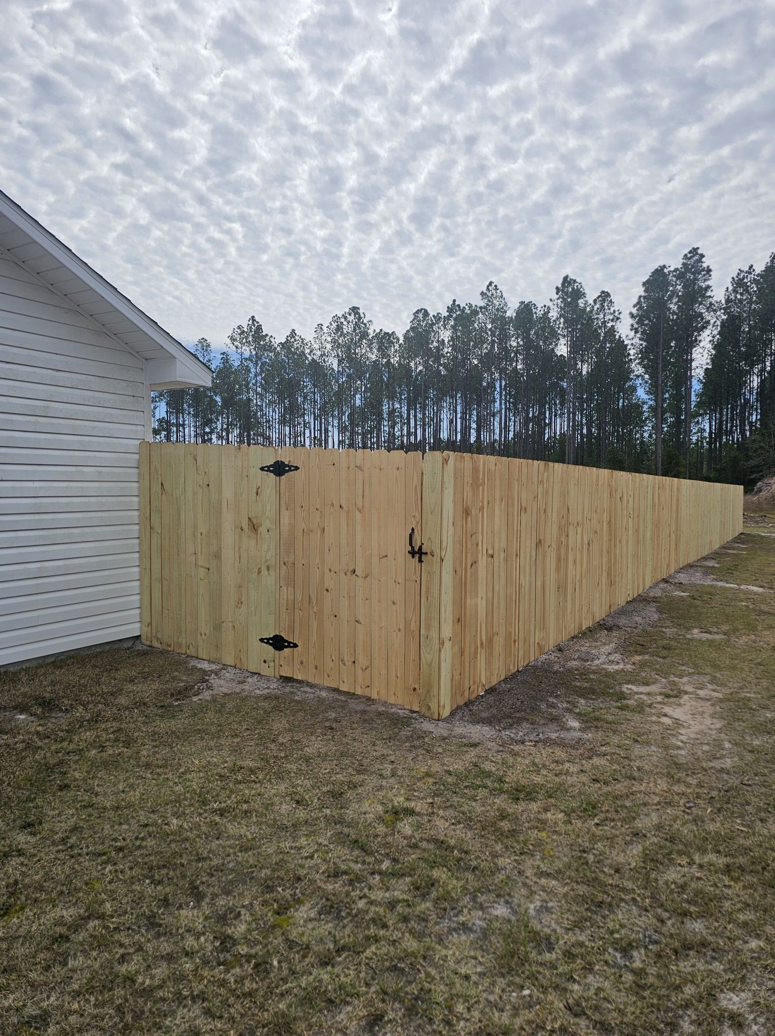 A newly installed light-wood privacy fence extends from the side of a white house toward a line of trees.