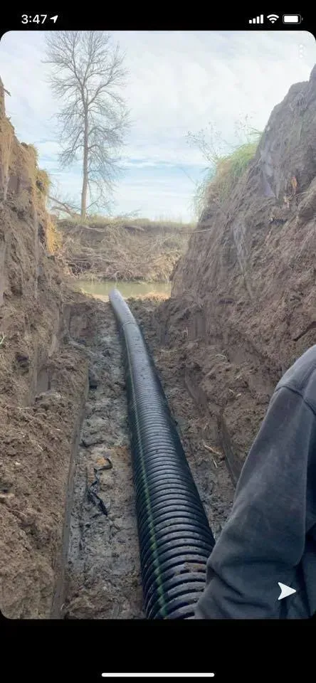 Black hose running through a narrow dirt trench on a grassy hillside under a bright sky