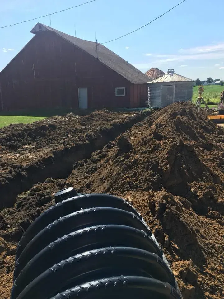 Farmyard with a large red barn, dirt mounds, and a black corrugated pipe in the foreground.