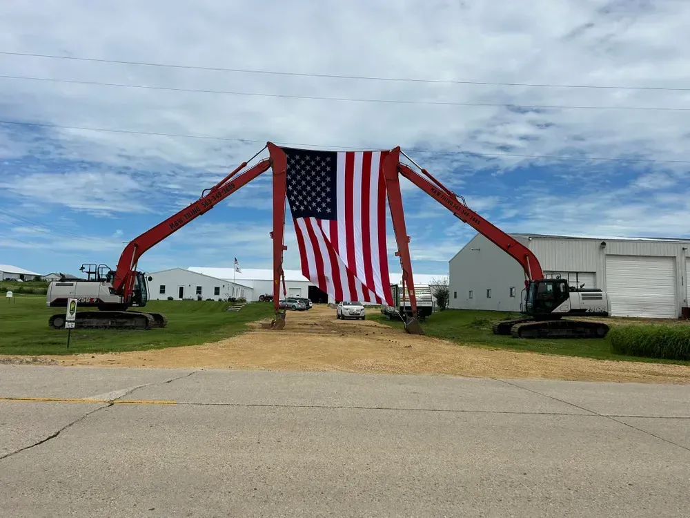 Two excavators lift a giant American flag between white buildings under a cloudy sky.