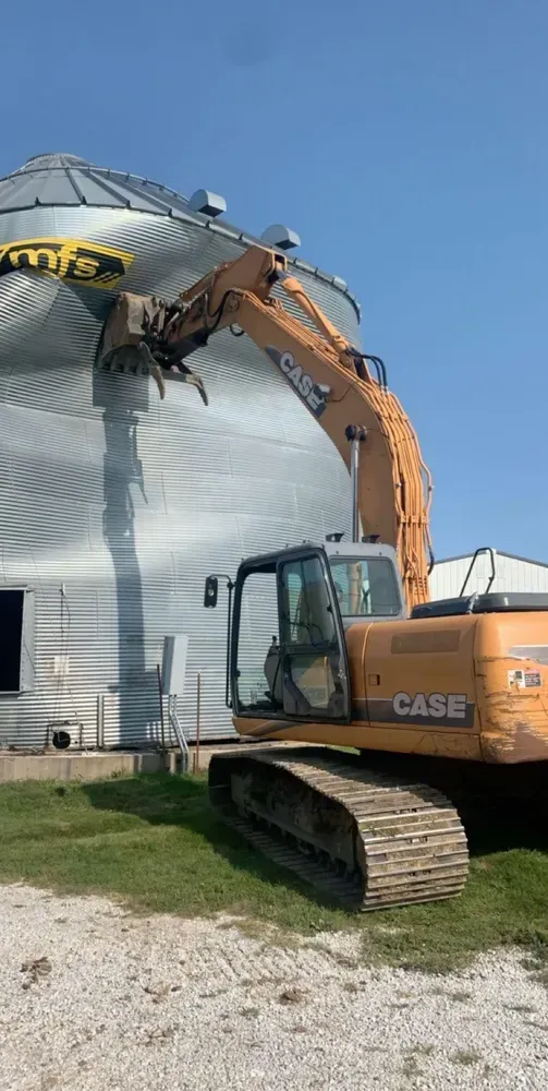 Orange excavator demolishing a metal grain silo under a blue sky