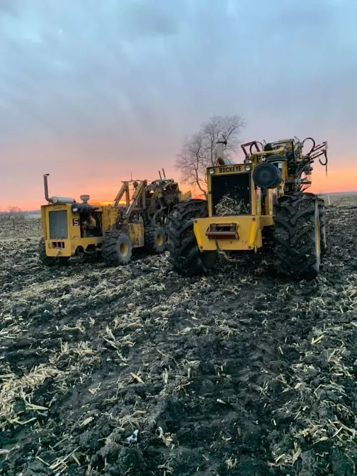 Yellow tractors in a muddy field at sunset with bare trees in the background