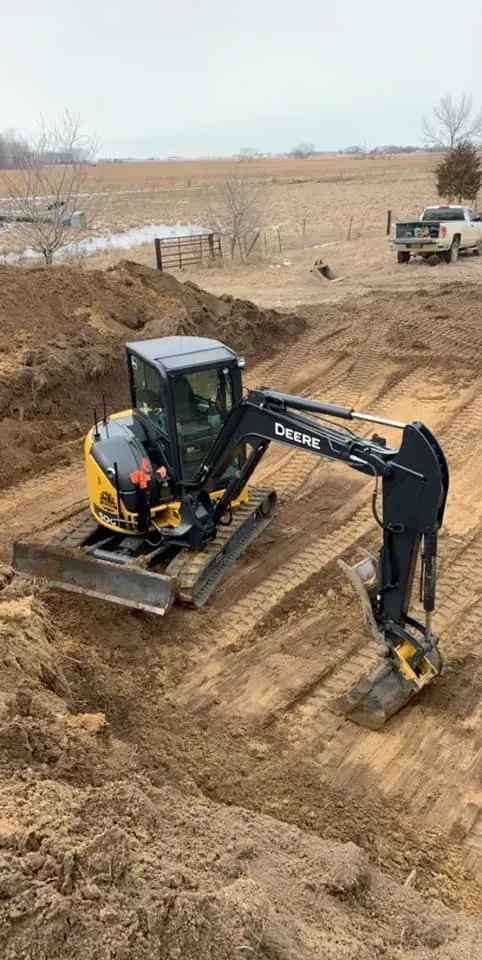 Excavator digging a sandy ditch at a construction site, with trucks and open fields in the background