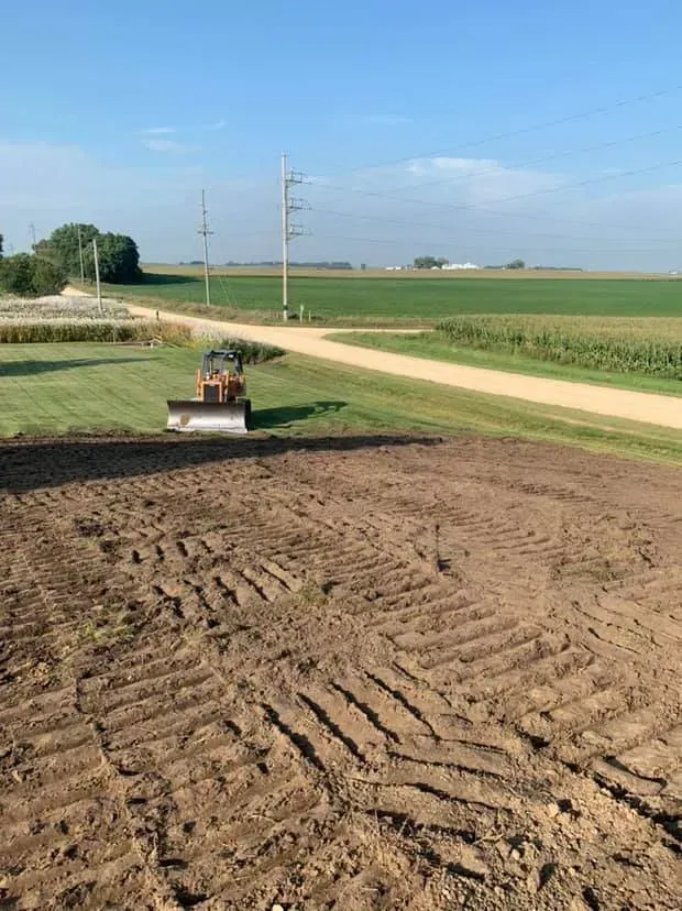 Freshly tilled field beside a rural road under a clear blue sky, with a small tractor in the distance