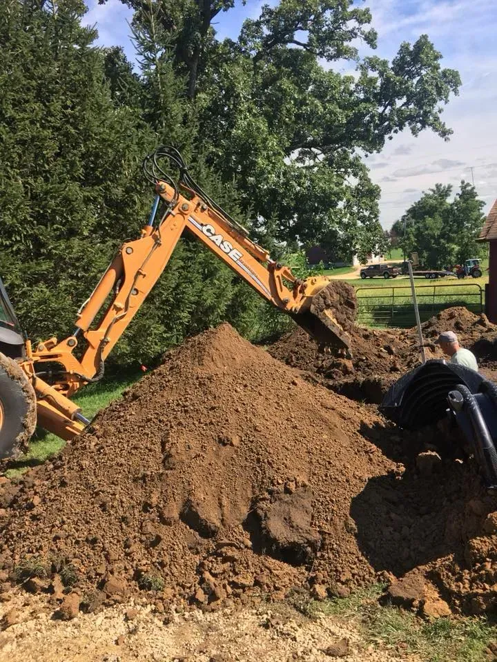 Orange excavator digging a large dirt pile while a worker crouches beside it in a grassy outdoor area.