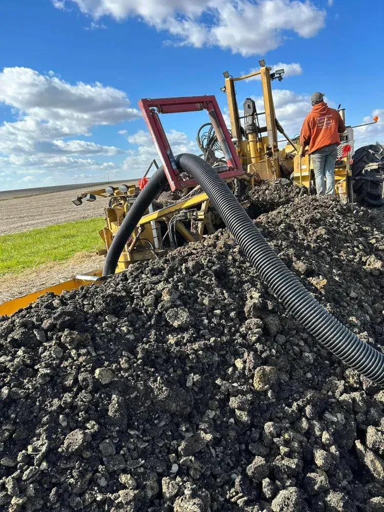 Construction workers by heavy machinery on a gravel mound under a cloudy blue sky