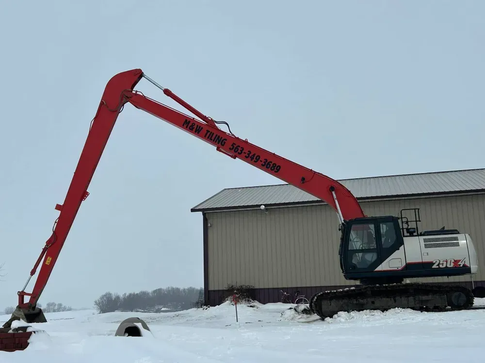 Red excavator extending over snow beside a metal building in a snowy field