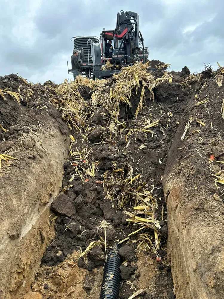 Excavator digging a trench through soil and crop debris under a cloudy sky