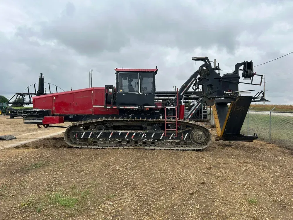 Red tracked farming machine with conveyor and trailer parked on a dirt lot under cloudy sky
