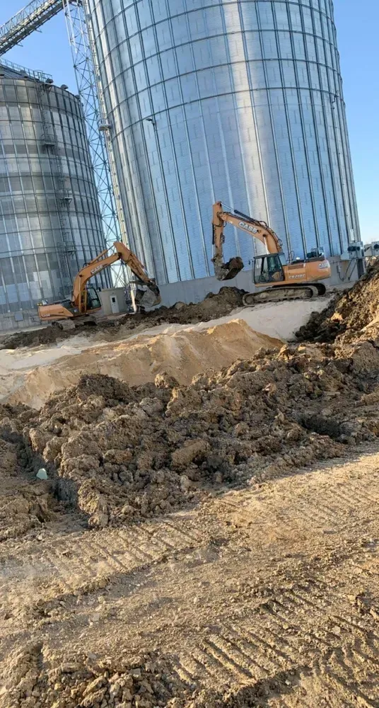 Excavators at a construction site beside tall glass silos, with dirt piles and earthmoving equipment.