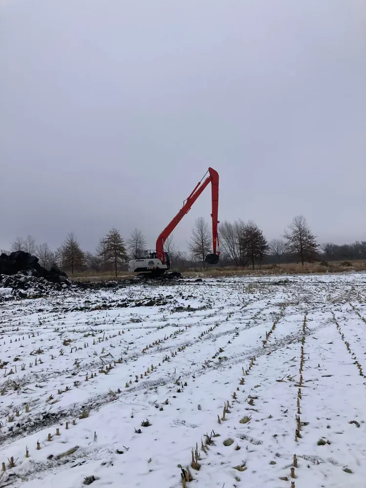 Red excavator arm in a snowy field under a gray sky, with bare trees in the background.
