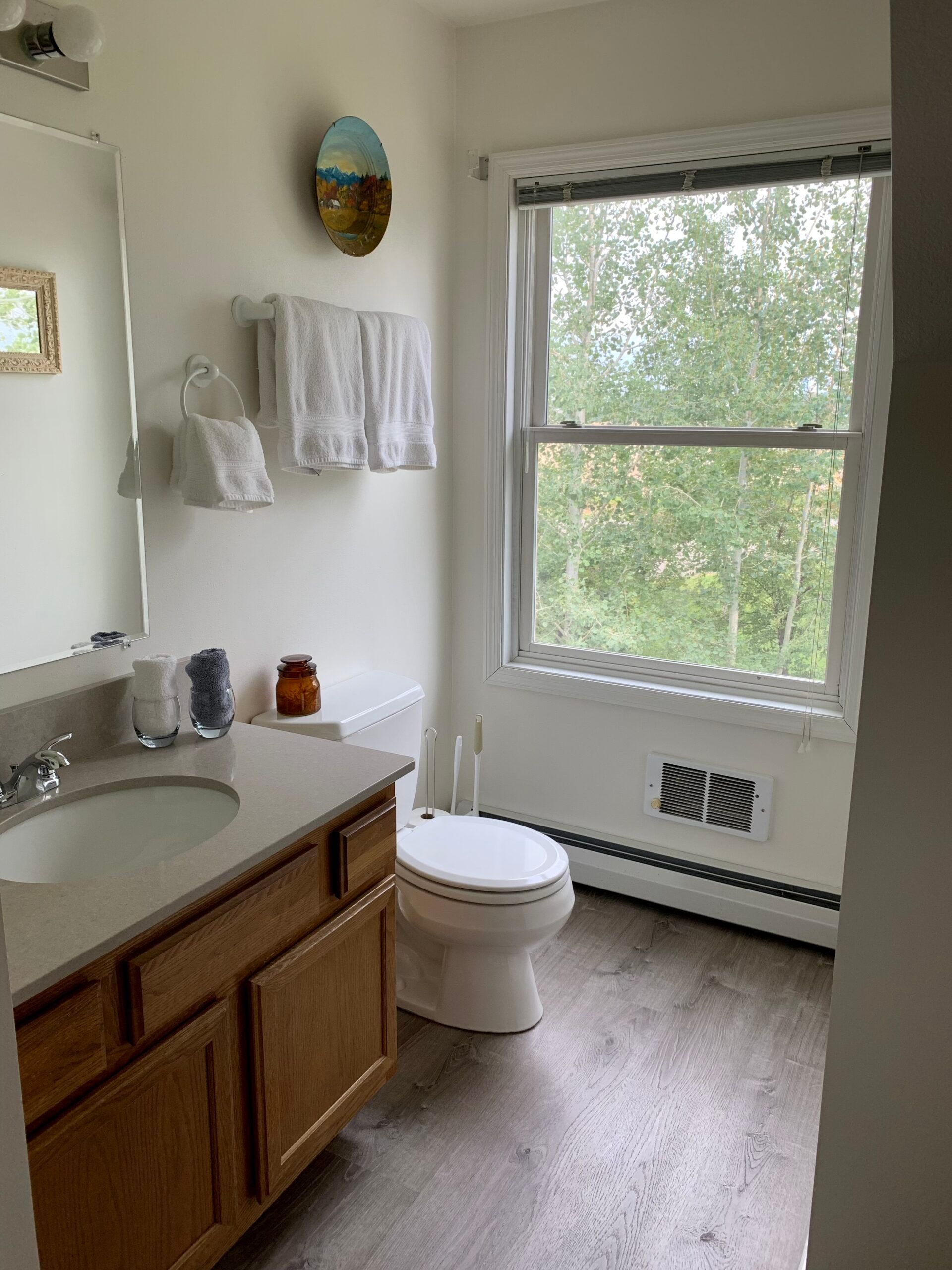 A bathroom with a toilet , sink , mirror and window.