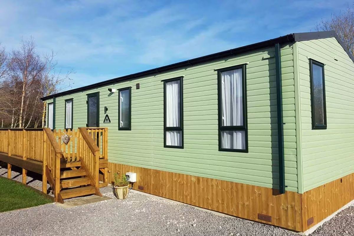 Green mobile home with wooden deck and stairs on a sunny day.