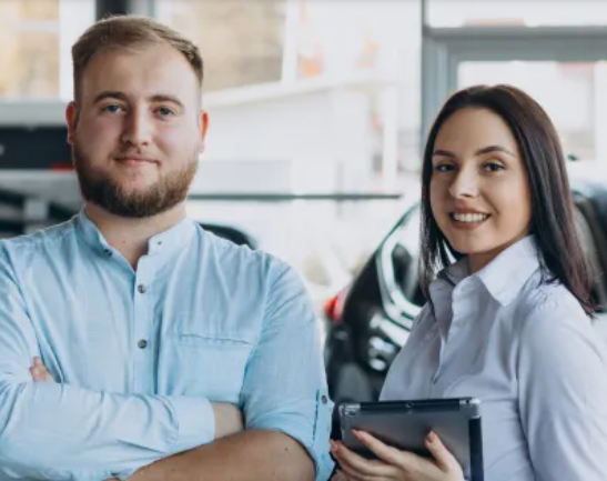 Dois vendedores de carros em uma concessionária. Um carro vermelho e preto é visível. A mulher segura um tablet.