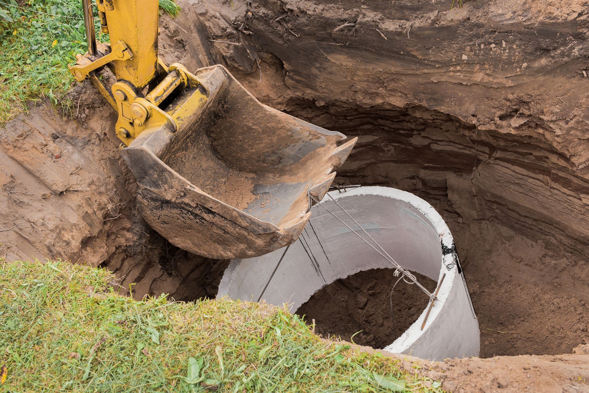 An excavator, equipped with a large bucket, is being lowered into the pit using sturdy steel cables. It is tasked with constructing a concrete sewer ring for a home culvert.