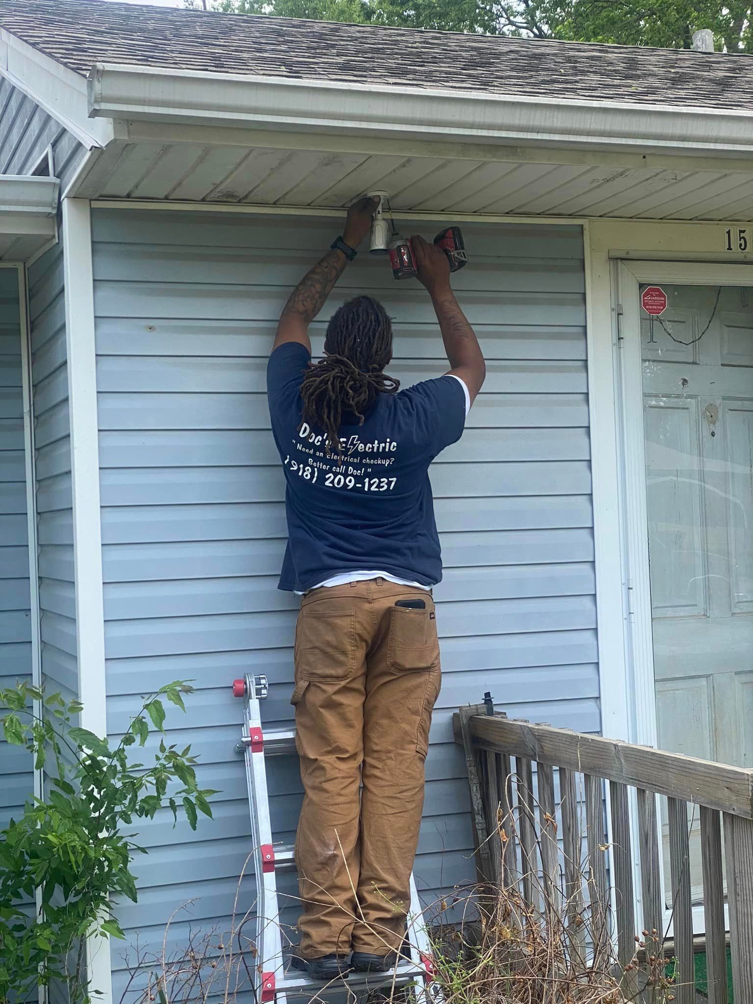 A man is standing on a ladder fixing a light on the side of a house.