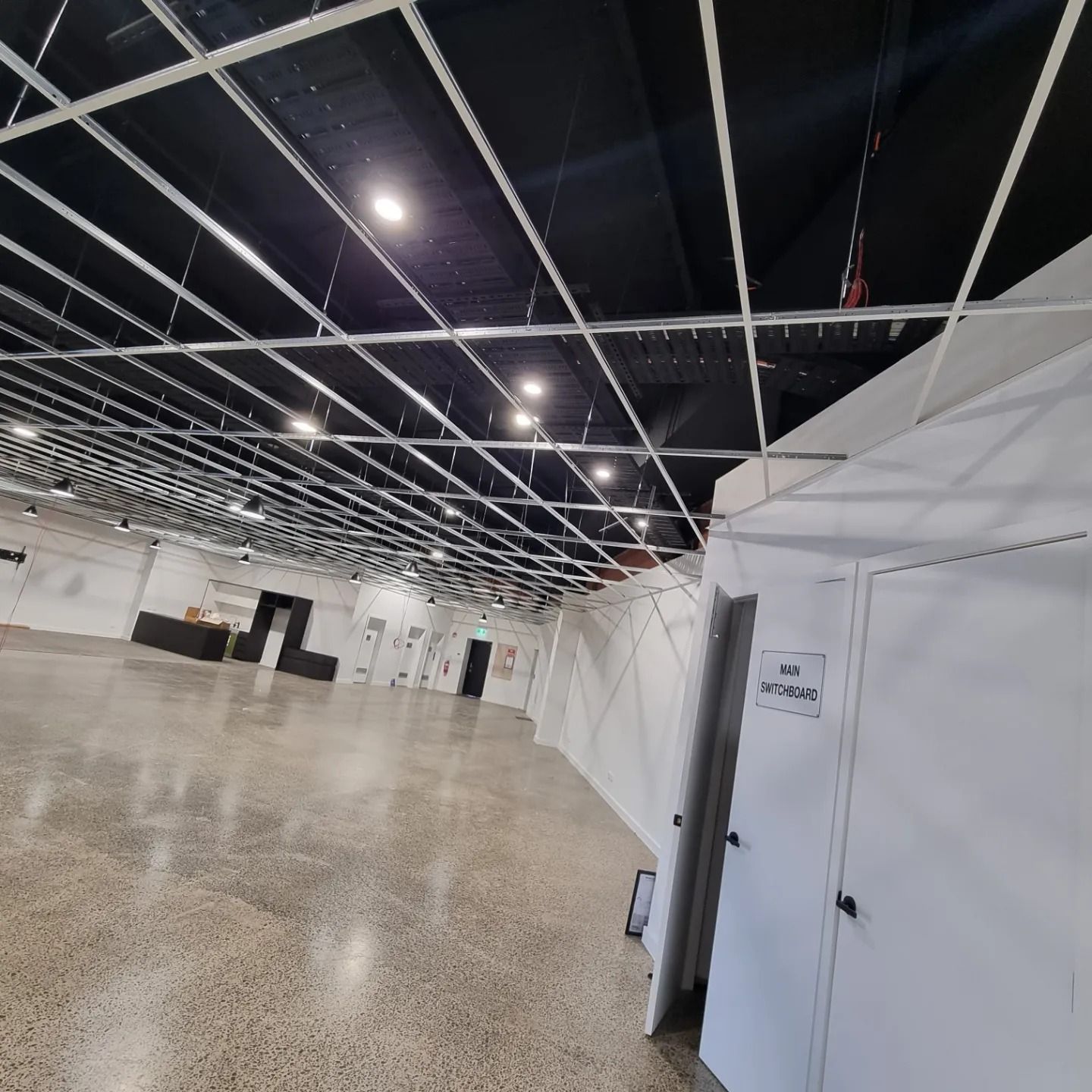 A large, empty hall with a concrete floor, a black ceiling, and an unfinished white metal grid frame hanging above.