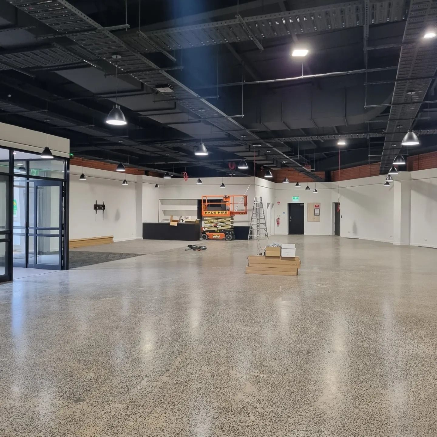 An empty, industrial-style room with white walls, polished concrete floors, and exposed dark ceilings with pendant lights.