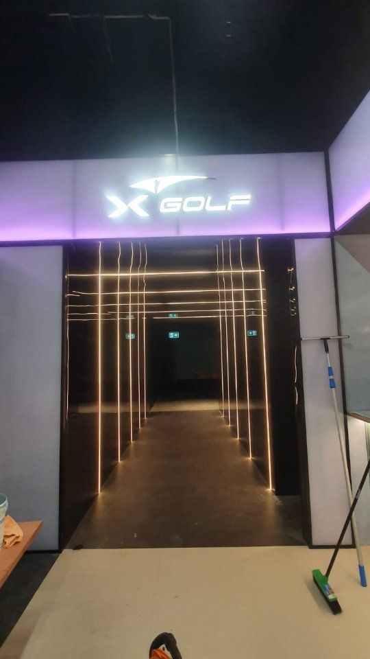 Entrance to an X-GOLF venue with a glowing sign above a dark hallway illuminated by thin vertical LED light strips.