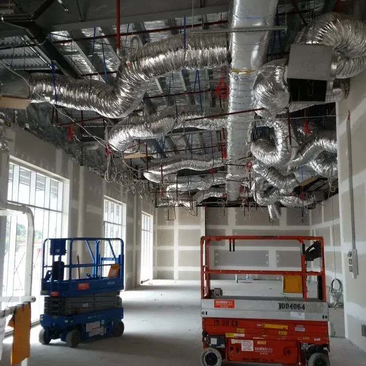 An indoor construction site features an exposed ceiling with silver ductwork, with blue and orange scissor lifts below.