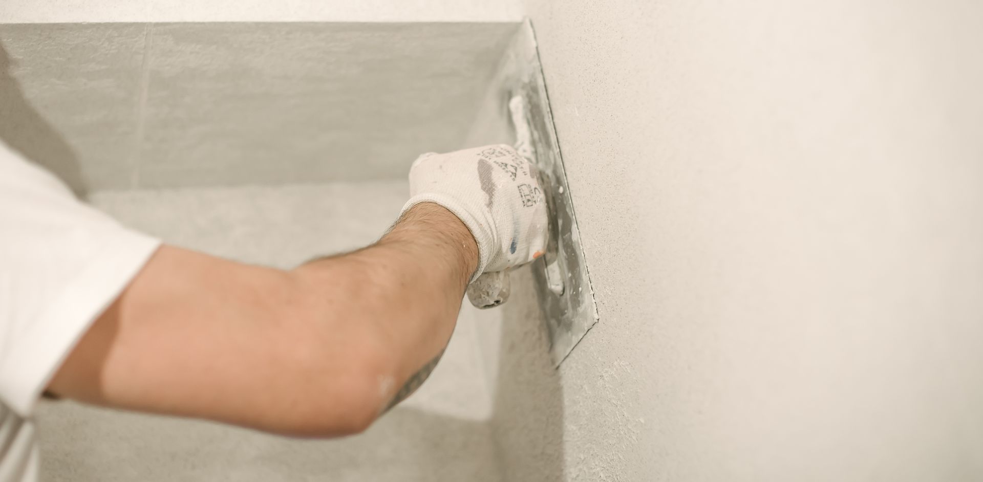 A person wearing a white glove uses a metal trowel to smooth plaster onto a light-colored wall.