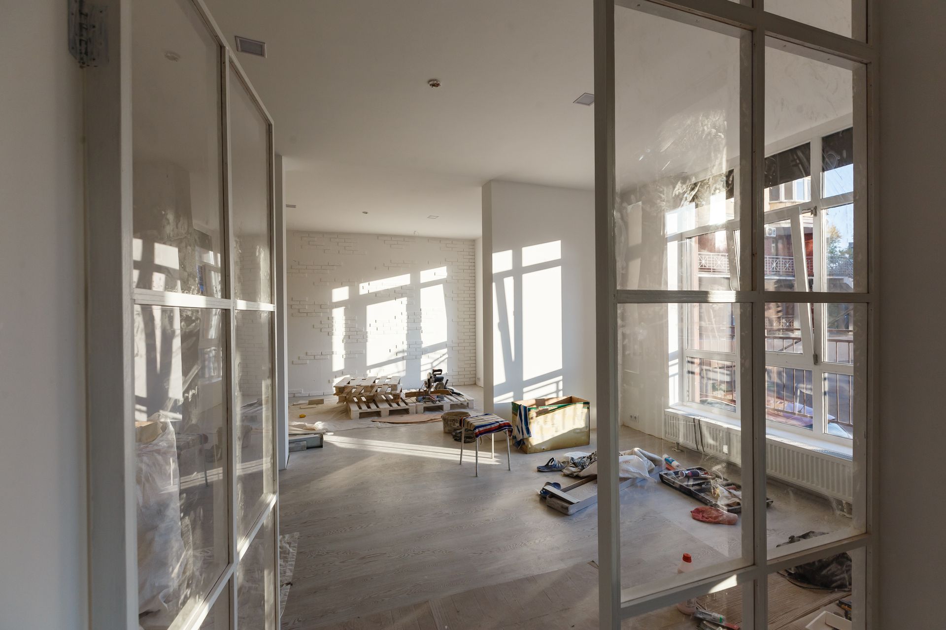 Interior of a bright, sunlit room under construction, viewed through a white, grid-patterned French glass door.