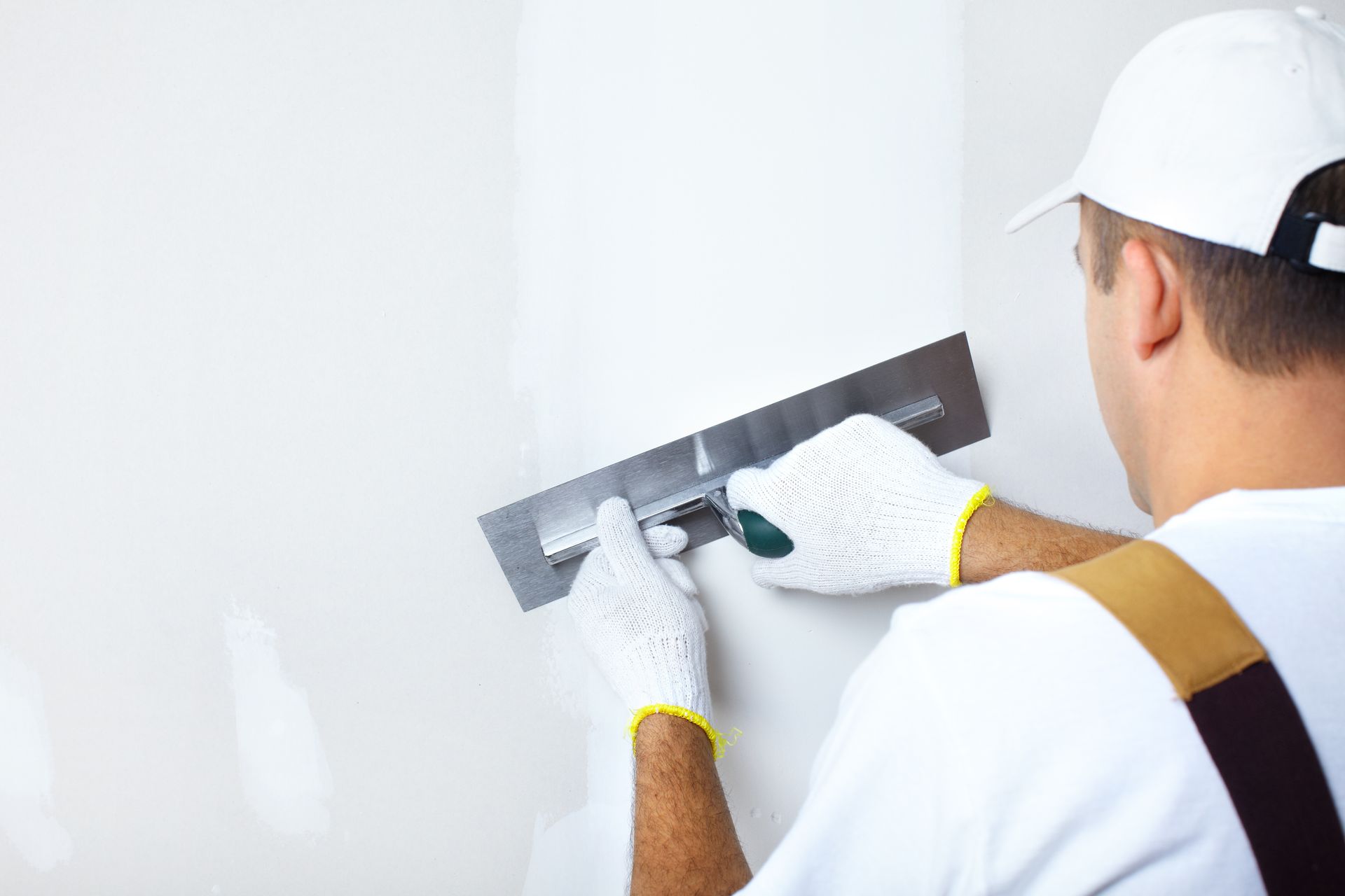 A worker in white workwear and gloves uses a metal trowel to smooth white plaster onto a wall.
