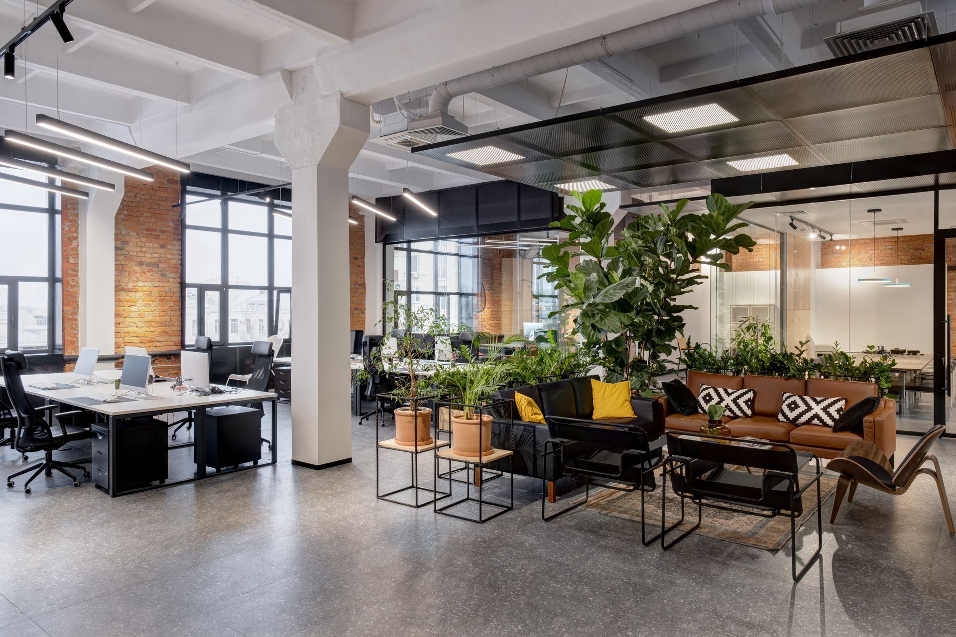 A modern, open-plan office with brick walls, exposed ceilings, desk workspaces, and a lounge area with plants.