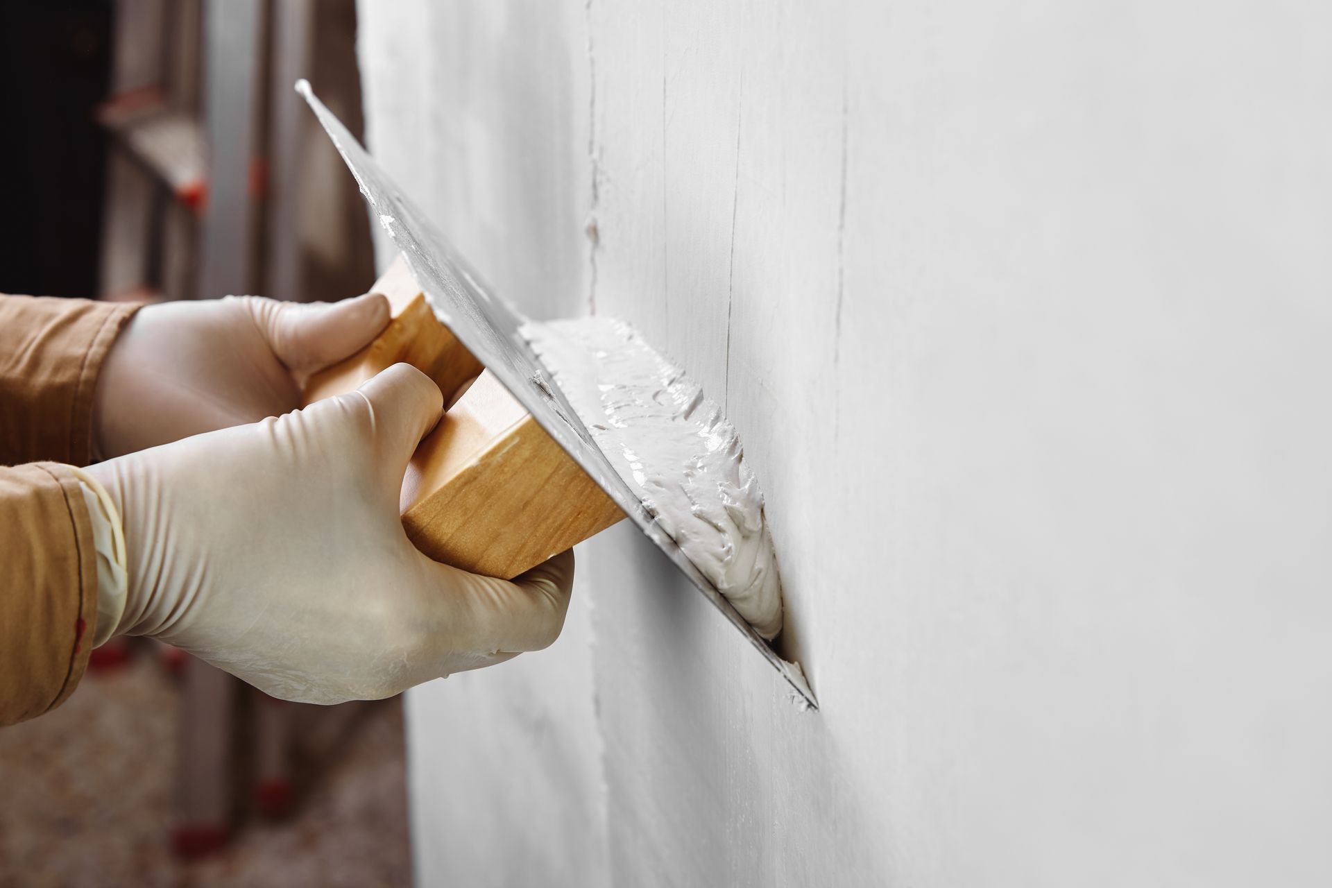 A gloved hand uses a metal trowel to smooth white joint compound onto a wall.