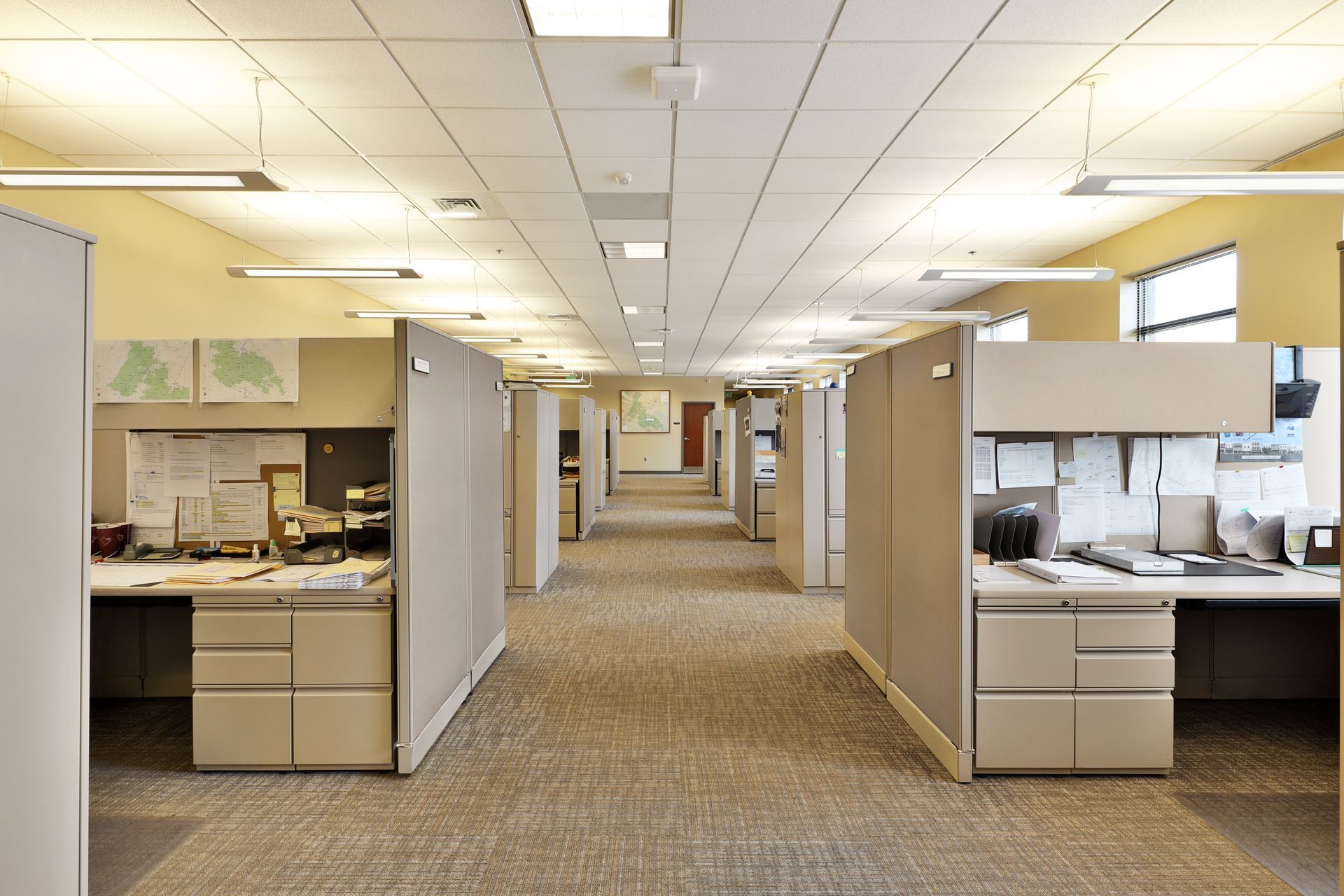 A bright, empty office space with rows of modular beige cubicles and desks leading toward a hallway and doorway.