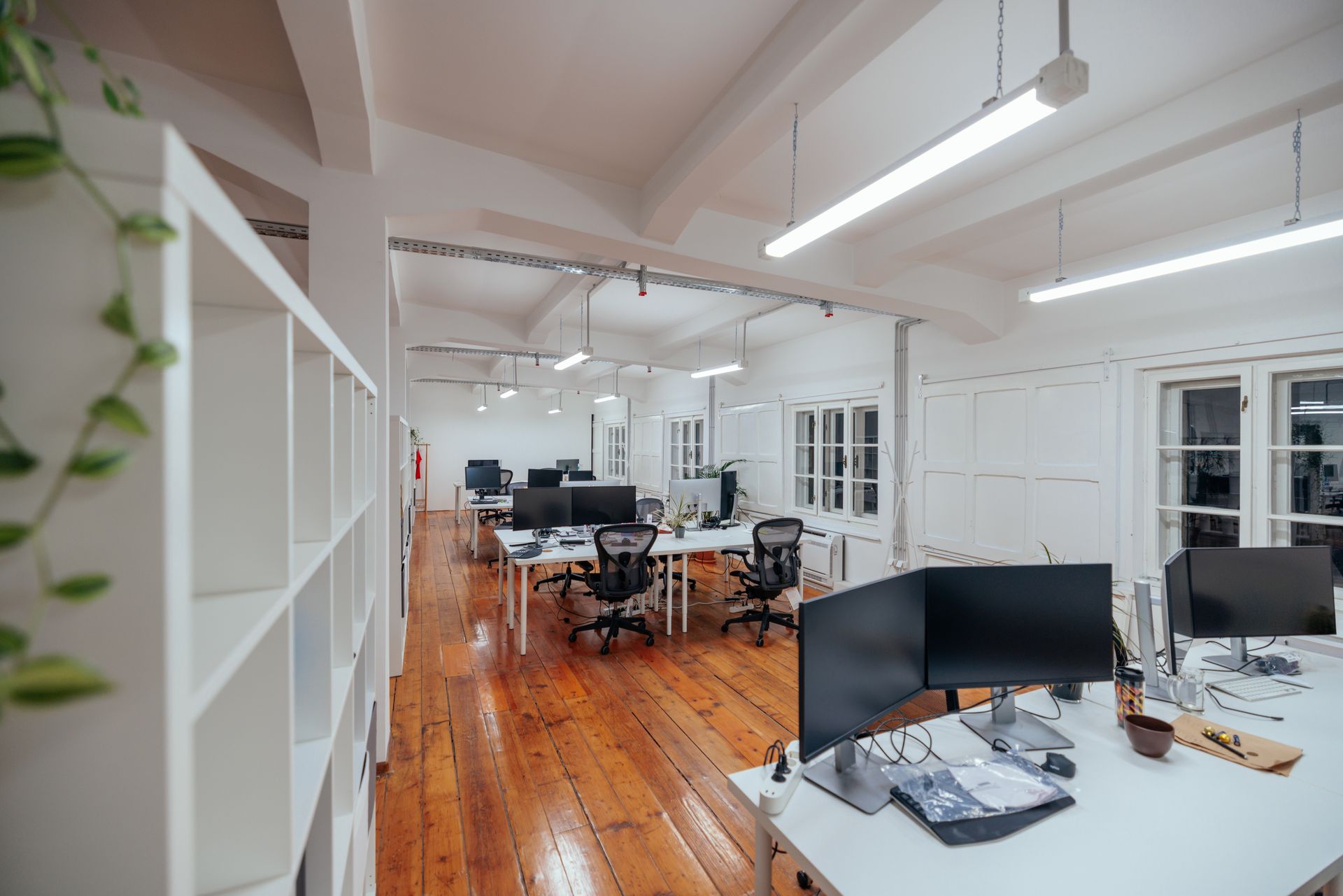 An office interior with white desks, black computer monitors, and chairs on a wood floor under long hanging lights.