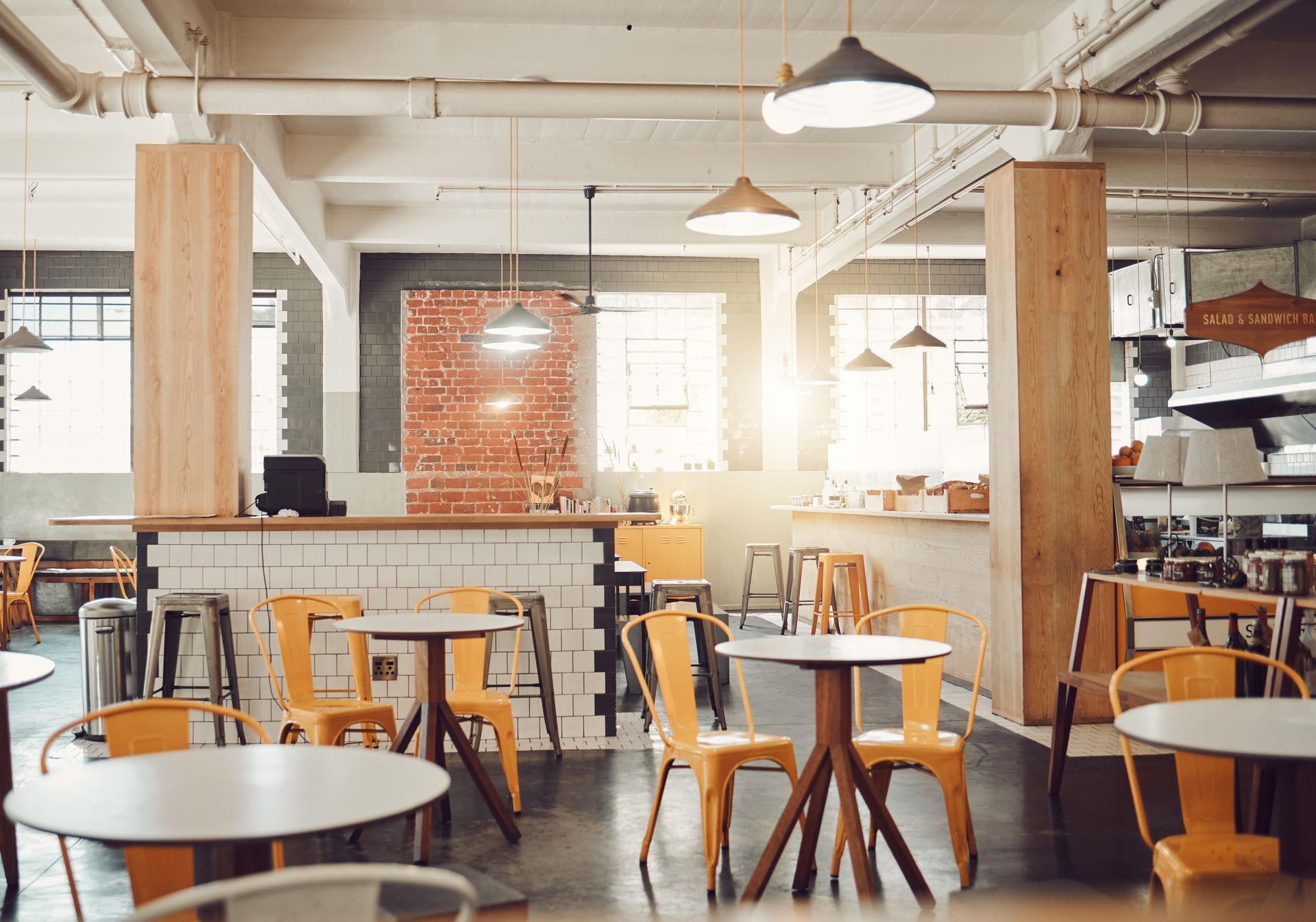 A modern cafe interior with industrial decor, wooden pillars, brick accents, and several round tables with yellow chairs.
