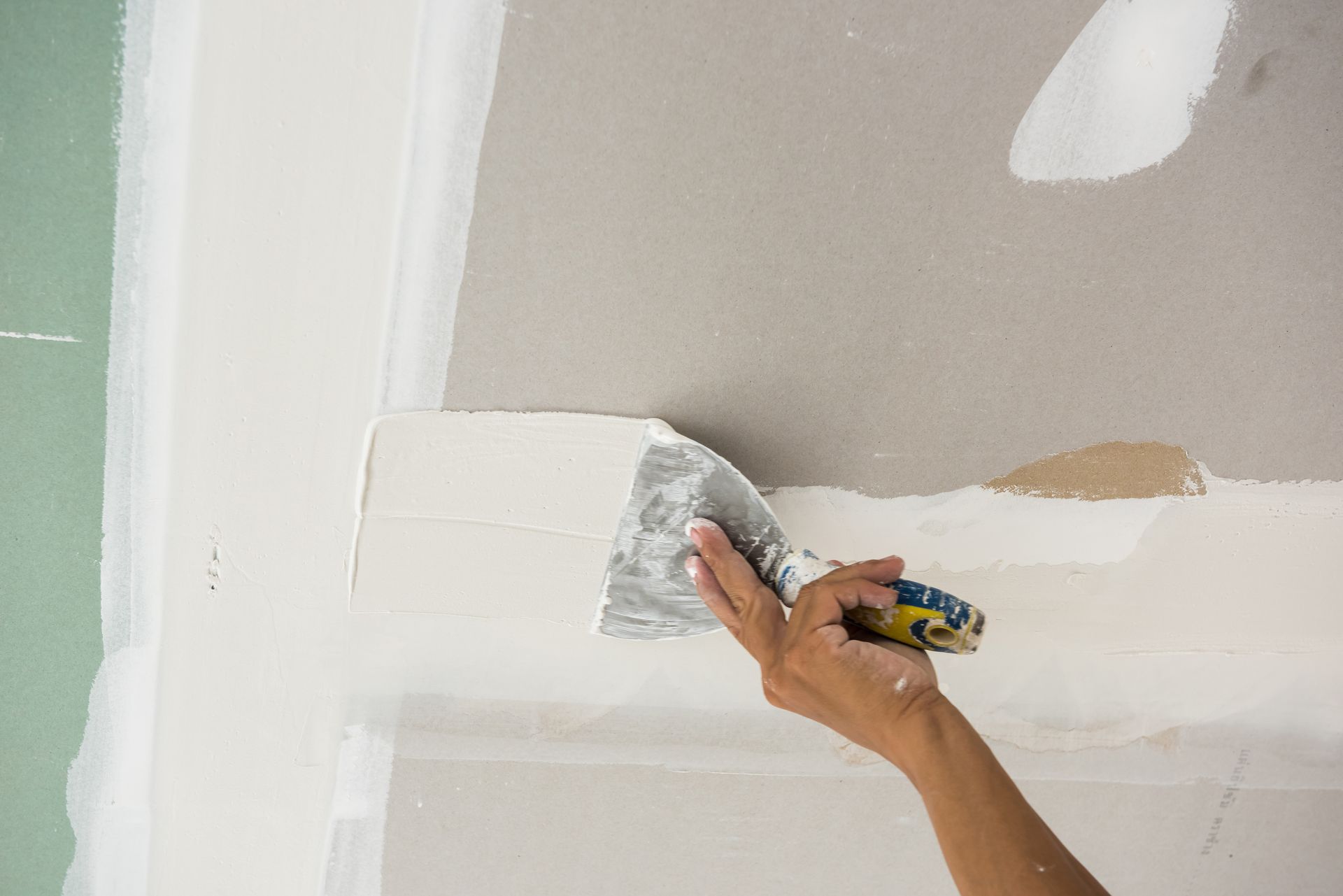 A hand uses a metal putty knife to spread joint compound over a seam on a drywall ceiling.