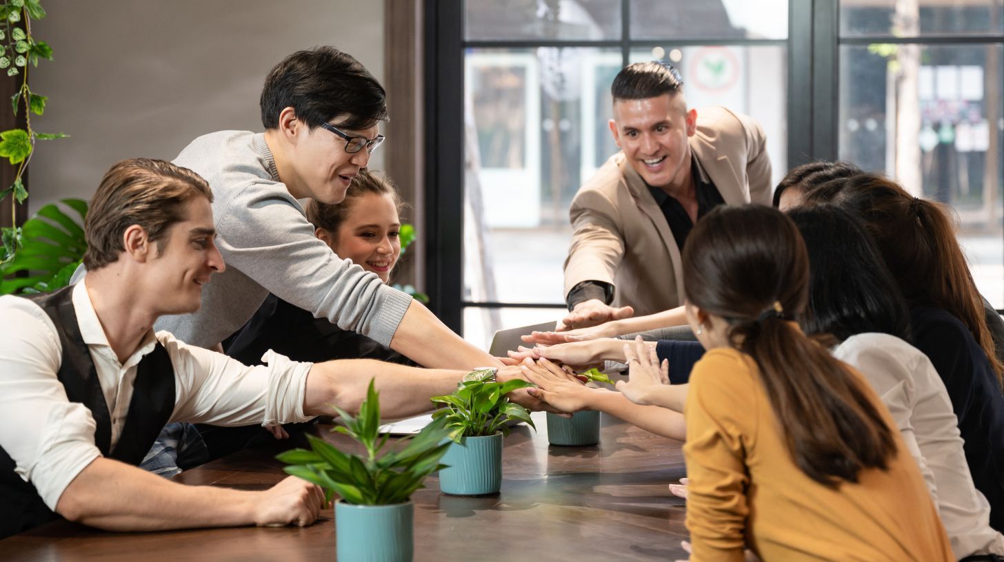 A diverse group of colleagues in a modern office gather around a wooden table, smiling as they stack their hands together.