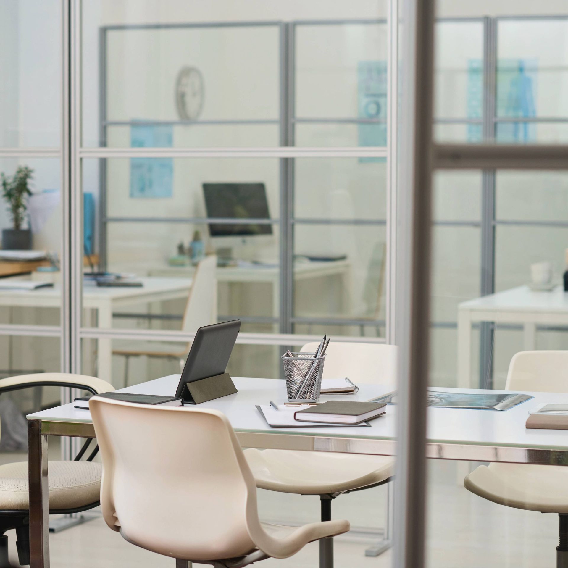 A modern office space with white desks, light-colored chairs, a tablet, and notebooks viewed through glass partitions.