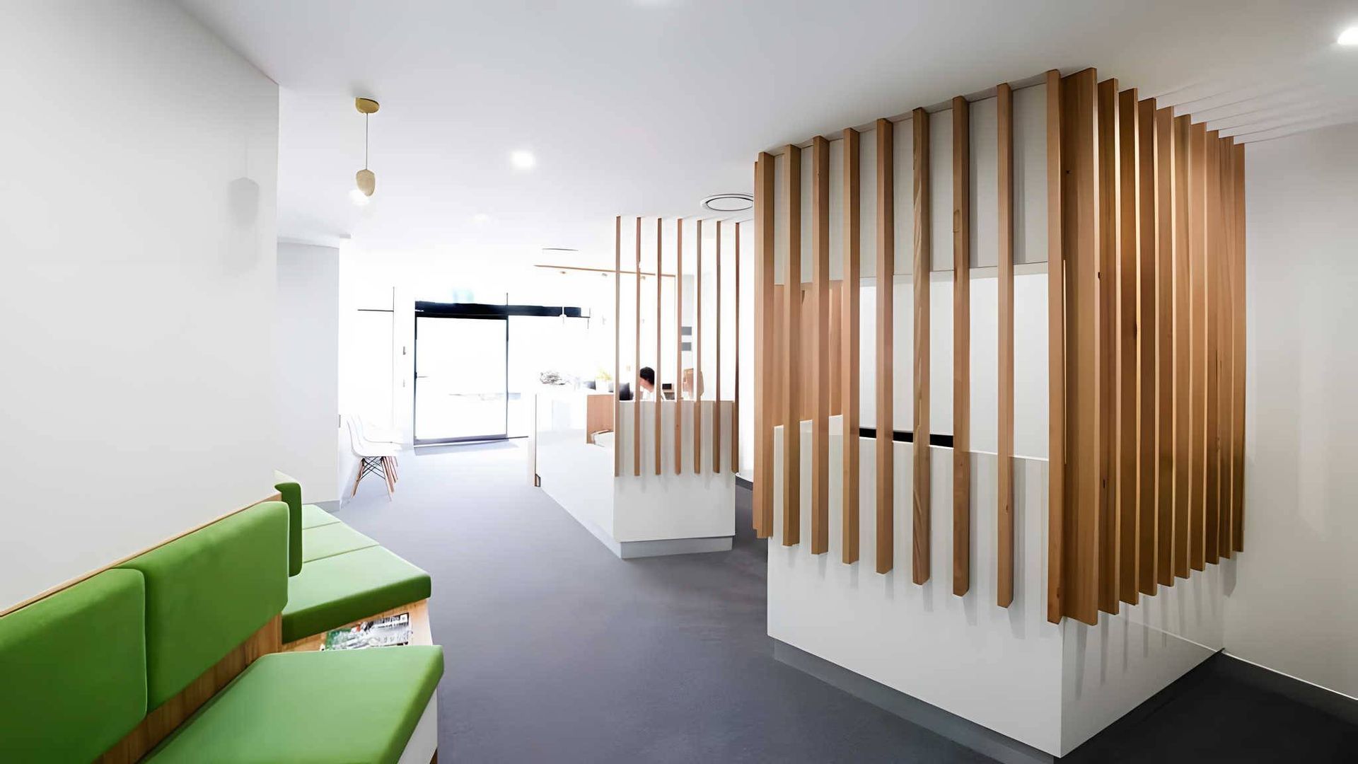 A modern waiting area with green bench seating, grey flooring, and a reception desk accented by vertical wood slats.
