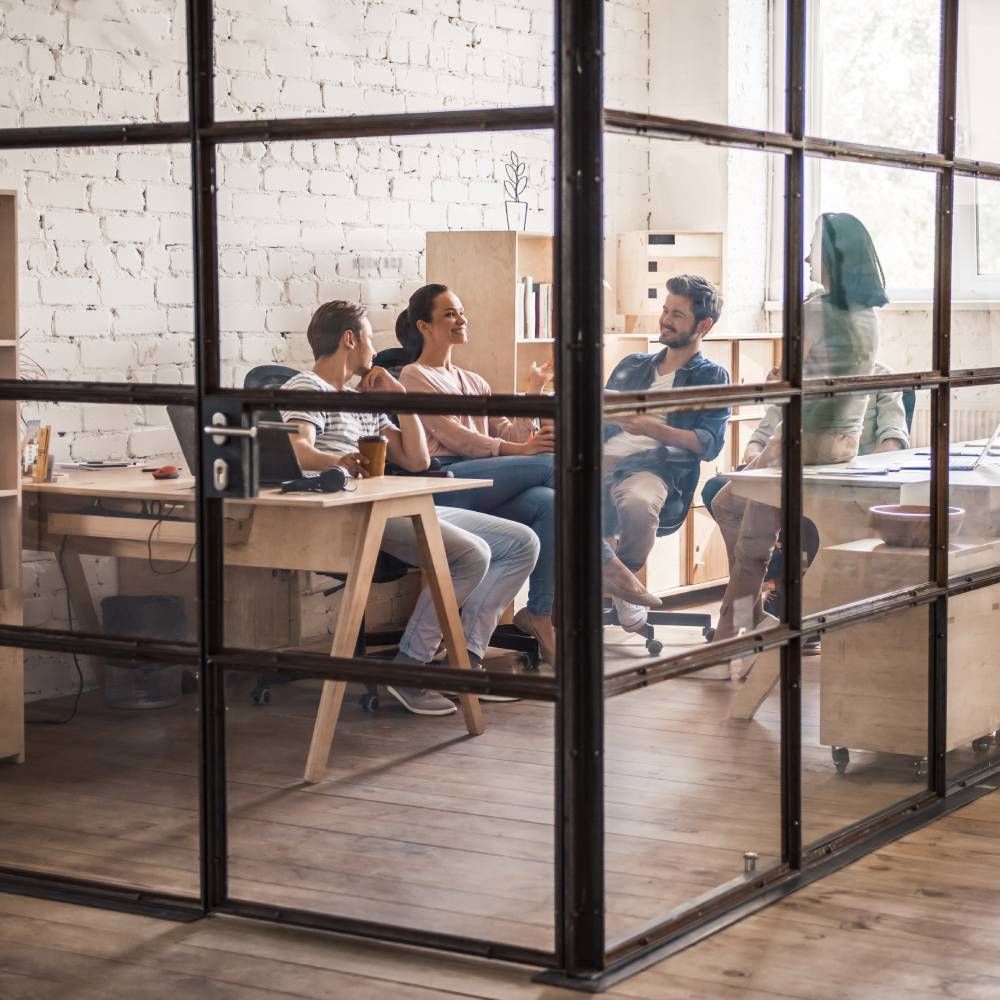 Four people collaborate in a modern, light-filled office with glass walls and white brick, engaged in a group discussion.