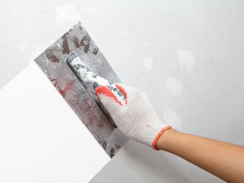 A hand in a white work glove uses a metal trowel to spread joint compound or plaster across a white wall.