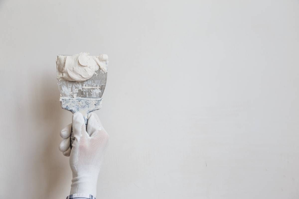 A hand in a white glove holds a putty knife covered in white spackle against a blank white wall.