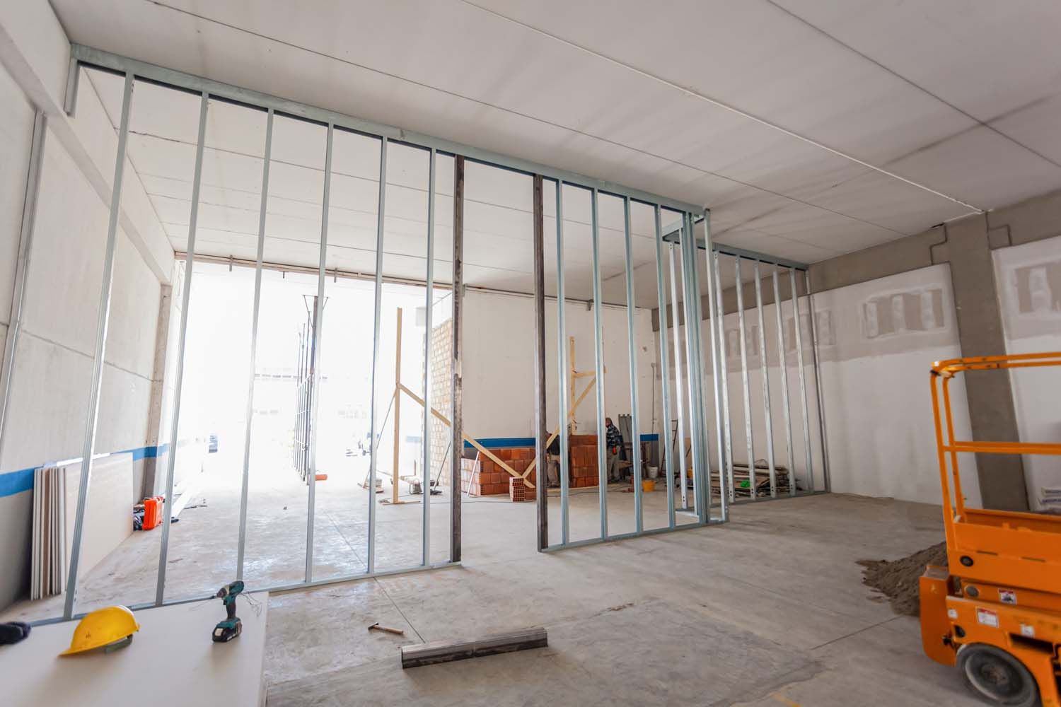 A metal stud frame for a wall being built inside an industrial space, with a yellow hard hat on a desk in the foreground.