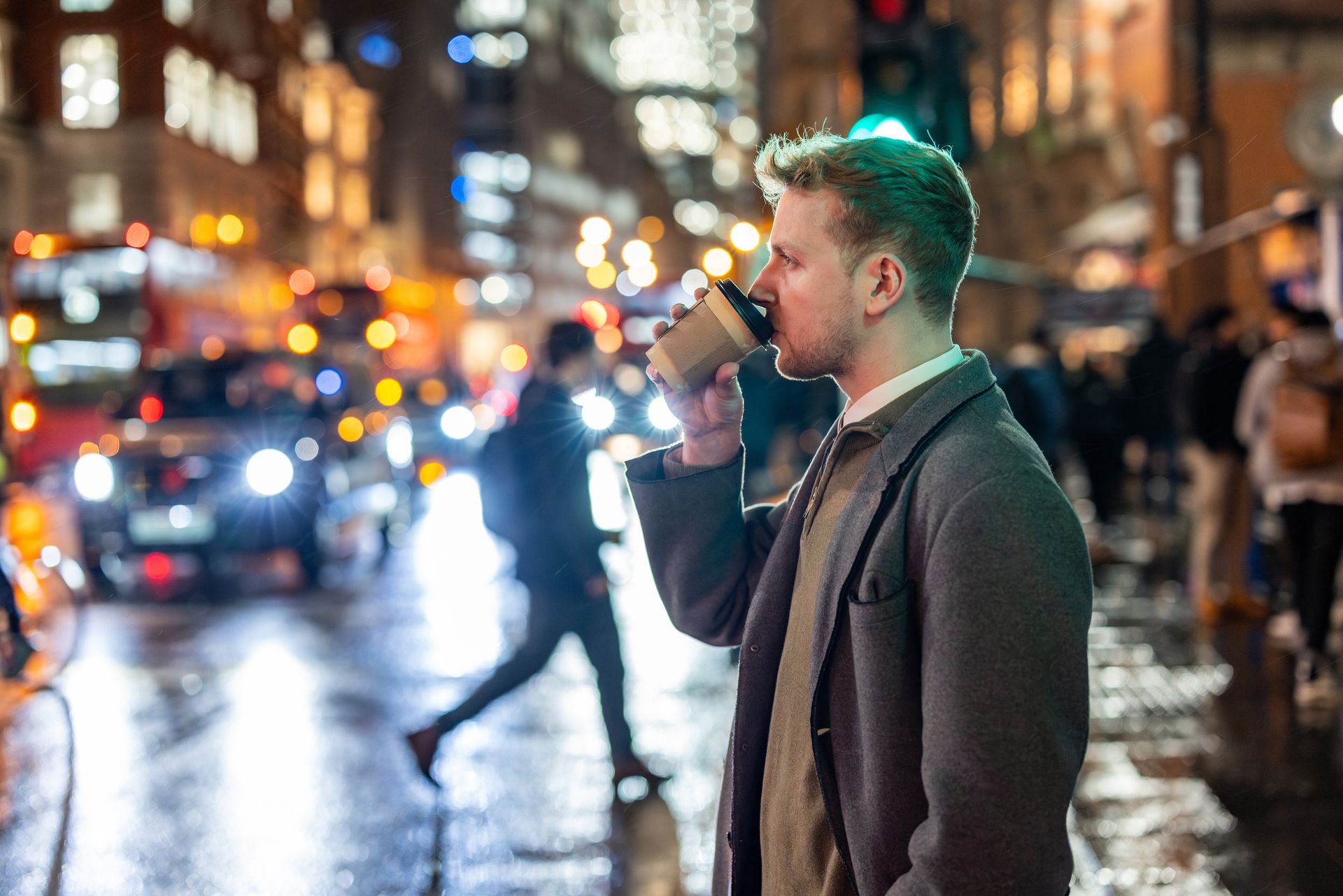 Man in a gray coat drinking from a cup on a rainy city street with blurred traffic lights at night