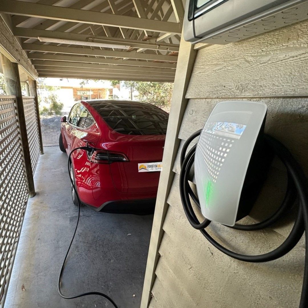 Red Tesla Charging Next to a White Charging Station on a House. Car is Under a Carport — Forster Solar and Lighting In Forster, NSW
