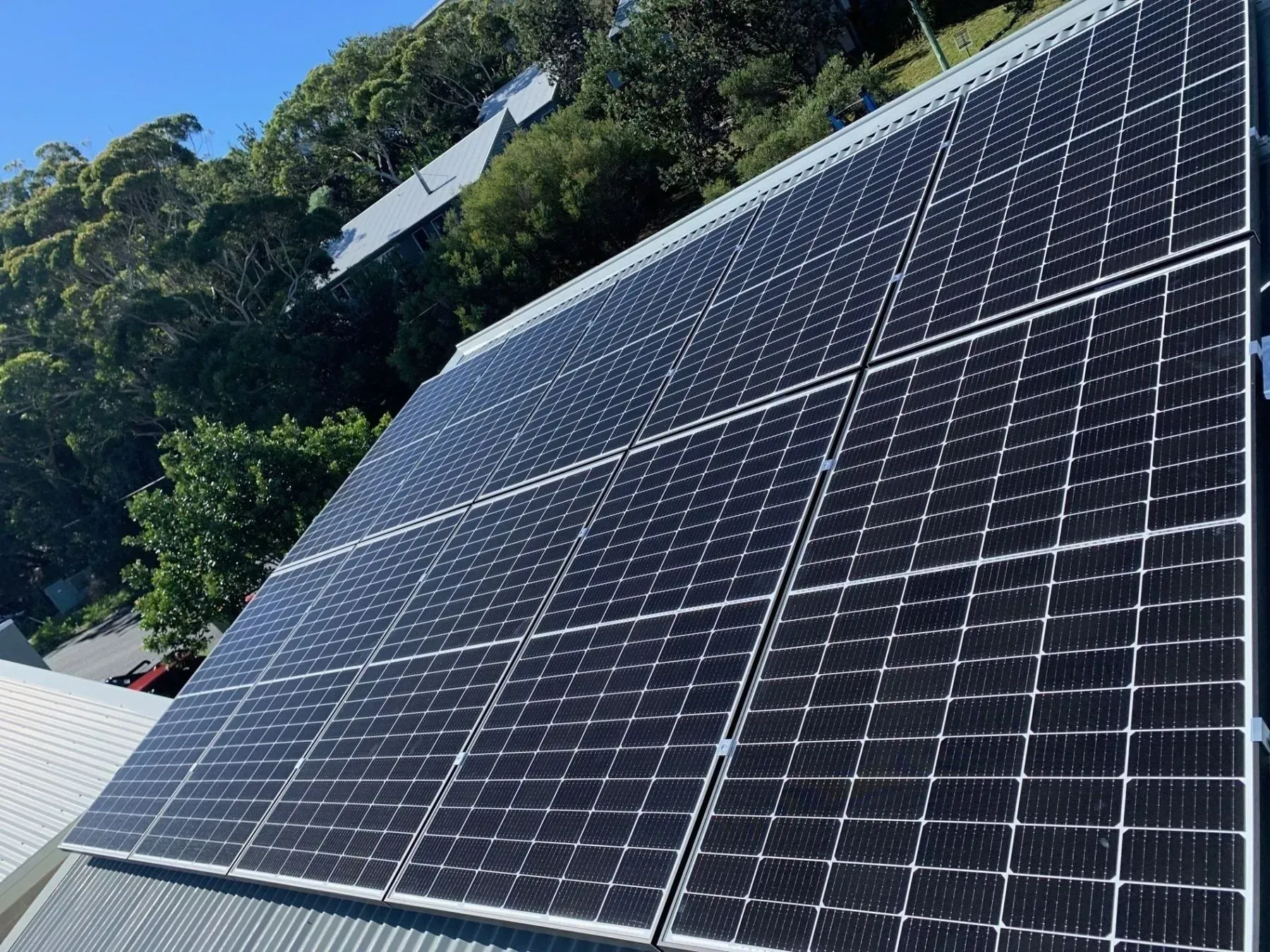 Solar Panels on a Rooftop, Angled Towards the Sun Trees and Buildings in the Background — Forster Solar and Lighting In Forster, NSW
