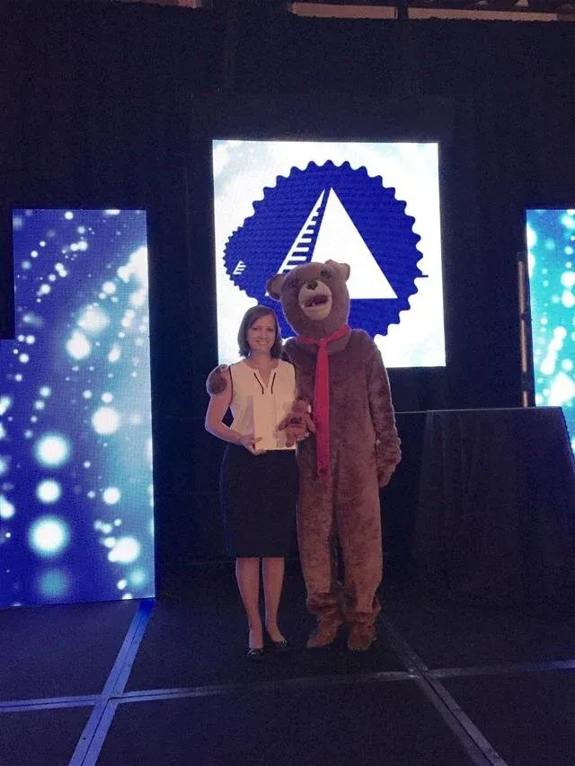 Woman in black skirt and white top with a bear mascot on stage. Blue backdrop with logo and lights.