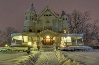 Snow-covered Victorian house at dusk, lit porch, two turrets, pathway with snow.