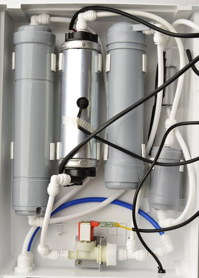 Vista interna di un'unità di filtrazione dell'acqua con tre contenitori filtranti verticali, tubi e una piccola valvola solenoide.