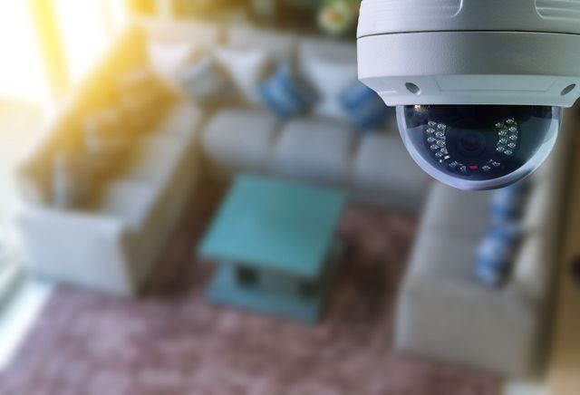 Una telecamera di sicurezza a cupola montata sul soffitto, che inquadra un soggiorno sfocato con divani e un tavolino da caffè.