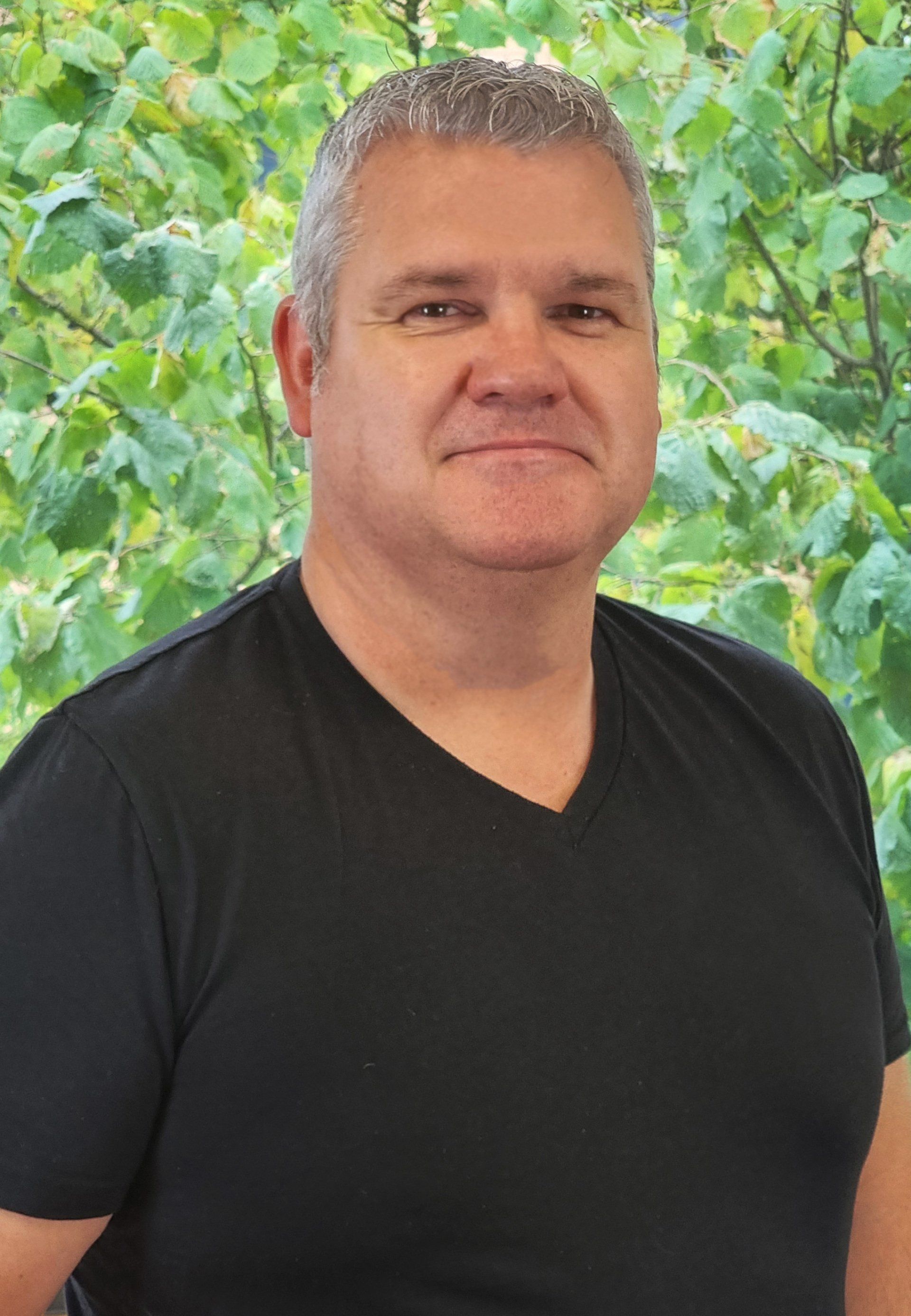 A man in a black shirt is standing in front of a tree.