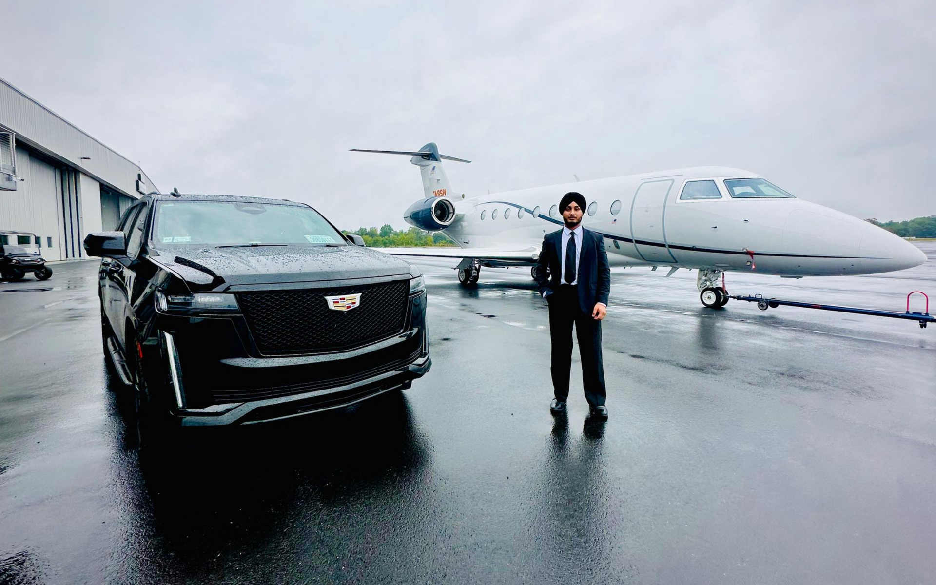 Man in a suit stands next to a black SUV and a private jet on a wet tarmac.