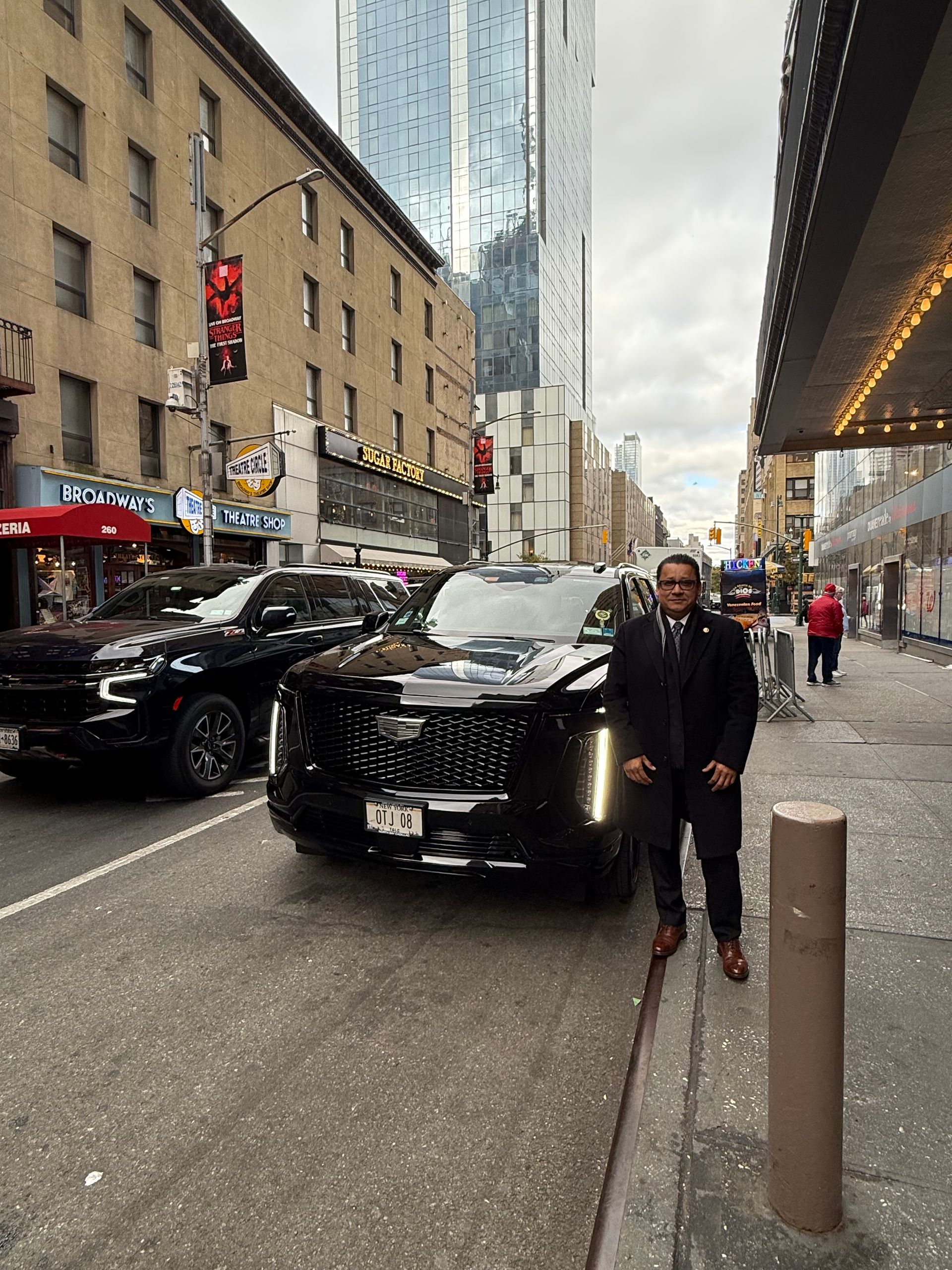 Man in black suit stands beside a black Cadillac Escalade on a city street; another black car behind it.
